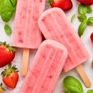 Honey strawberry basil popsicles on wooden sticks with fresh strawberries and basil leaves on a white wood background.