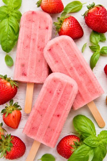 Honey strawberry basil popsicles on wooden sticks with fresh strawberries and basil leaves on a white wood background.