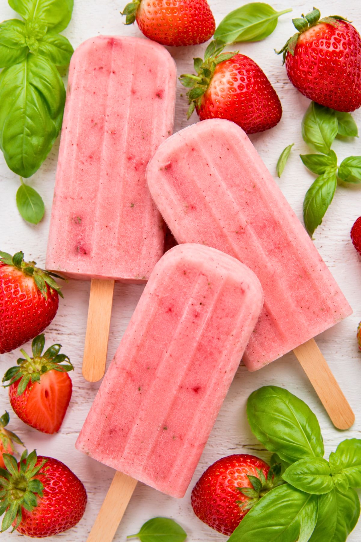 Honey strawberry basil popsicles on wooden sticks with fresh strawberries and basil leaves on a white wood background.