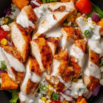 Close-up of a high-protein hot honey chicken sweet potato bowl with sliced chicken breast, roasted sweet potatoes, corn, tomatoes, lettuce, red onion, crumbled cheese, and creamy dressing.