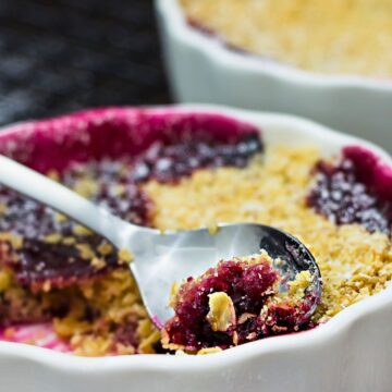 Individual mixed berry crumble in a white ramekin with golden oat topping and dark berry filling, served with a spoon on a black cooling rack.