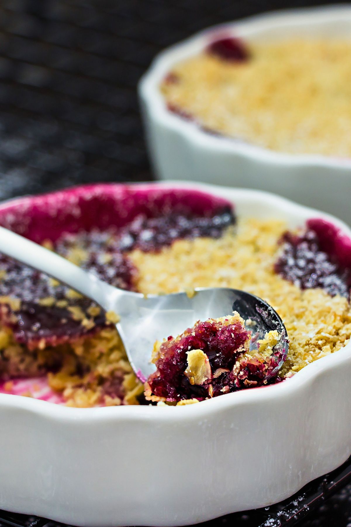 Individual mixed berry crumble in a white ramekin with golden oat topping and dark berry filling, served with a spoon on a black cooling rack.