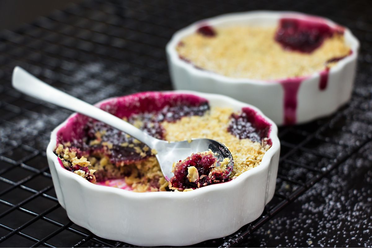 Individual mixed berry crumbles in white ramekins with golden oat topping and dark berry filling, served with a spoon on a black cooling rack.