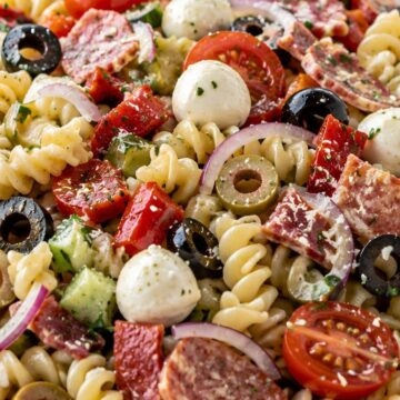 Italian antipasto pasta salad with rotini, salami, mozzarella balls, cherry tomatoes, cucumbers, red onion, black and green olives, red peppers, and herbs in a white bowl.