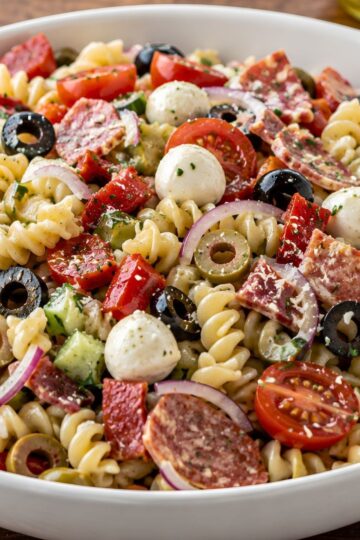 Italian antipasto pasta salad with rotini, salami, mozzarella balls, cherry tomatoes, cucumbers, red onion, black and green olives, red peppers, and herbs in a white bowl.