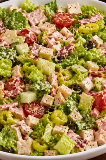 Italian grinder chopped salad with romaine lettuce, deli meat, provolone cubes, cucumber, cherry tomatoes, banana peppers, black olives, and creamy dressing in a white bowl.