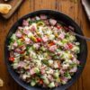 Italian grinder chopped salad with turkey, ham, salami, provolone, chopped vegetables, banana peppers, and garlic pita chips on a rustic wooden table.