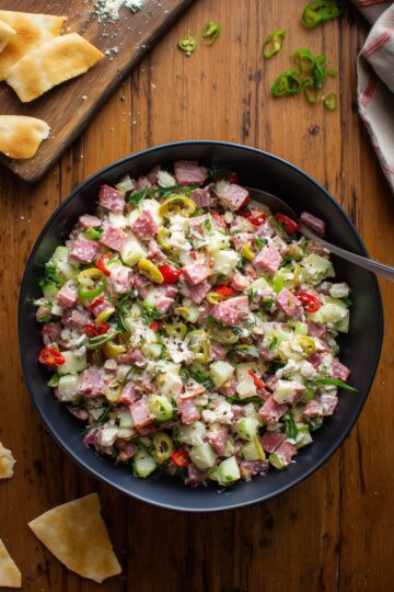 Italian grinder chopped salad with turkey, ham, salami, provolone, chopped vegetables, banana peppers, and garlic pita chips on a rustic wooden table.