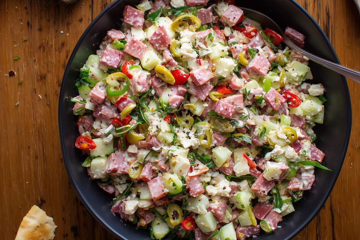 Italian grinder chopped salad with diced turkey, ham, salami, provolone, cucumbers, tomatoes, banana peppers, and lettuce in a black bowl.