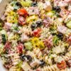 Italian grinder pasta salad with rotini, ham, salami, provolone, cherry tomatoes, cucumber, romaine, black olives, banana peppers, herbs, and creamy dressing in a white bowl.
