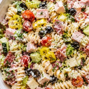 Italian grinder pasta salad with rotini, ham, salami, provolone, cherry tomatoes, cucumber, romaine, black olives, banana peppers, herbs, and creamy dressing in a white bowl.