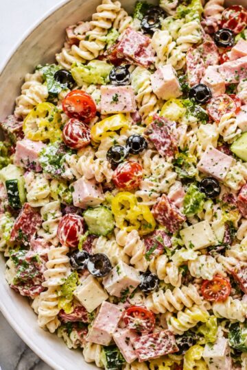 Italian grinder pasta salad with rotini, ham, salami, provolone, cherry tomatoes, cucumber, romaine, black olives, banana peppers, herbs, and creamy dressing in a white bowl.
