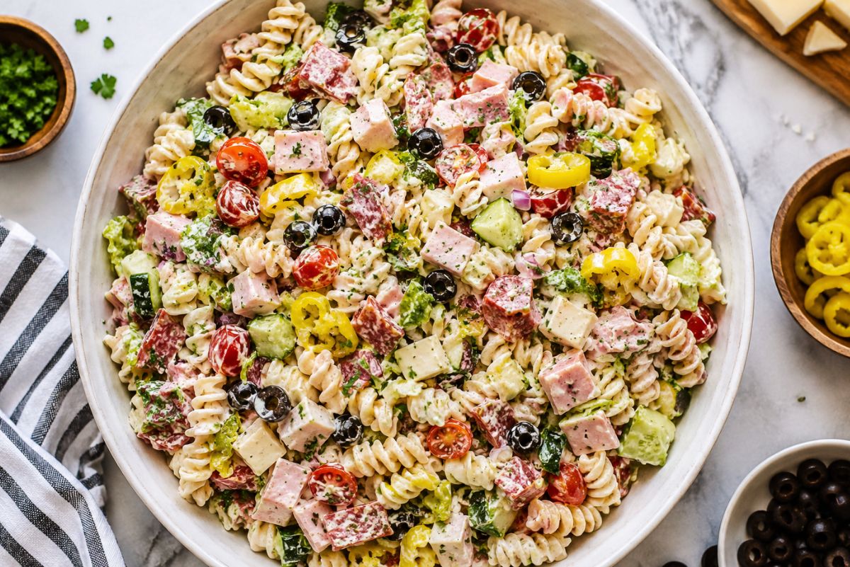 Italian grinder pasta salad in a large white bowl with rotini, ham, salami, provolone, cherry tomatoes, cucumber, romaine, black olives, banana peppers, parsley, and creamy dressing on a marble countertop.