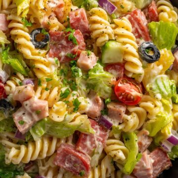 Close-up of Italian grinder pasta salad with rotini, salami, ham, lettuce, tomatoes, cucumbers, red onion, black olives, and creamy dressing in a black bowl.