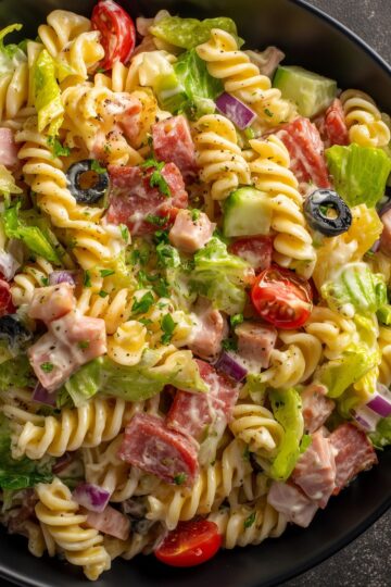 Close-up of Italian grinder pasta salad with rotini, salami, ham, lettuce, tomatoes, cucumbers, red onion, black olives, and creamy dressing in a black bowl.