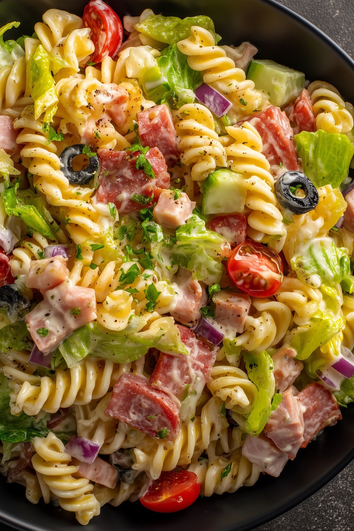 Close-up of Italian grinder pasta salad with rotini, salami, ham, lettuce, tomatoes, cucumbers, red onion, black olives, and creamy dressing in a black bowl.