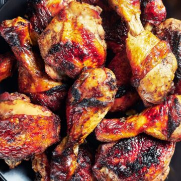 Jamaican jerk chicken drumsticks and thighs with charred, glossy skin piled on a black plate over a dark wood background.