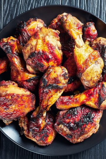 Jamaican jerk chicken drumsticks and thighs with charred, glossy skin piled on a black plate over a dark wood background.