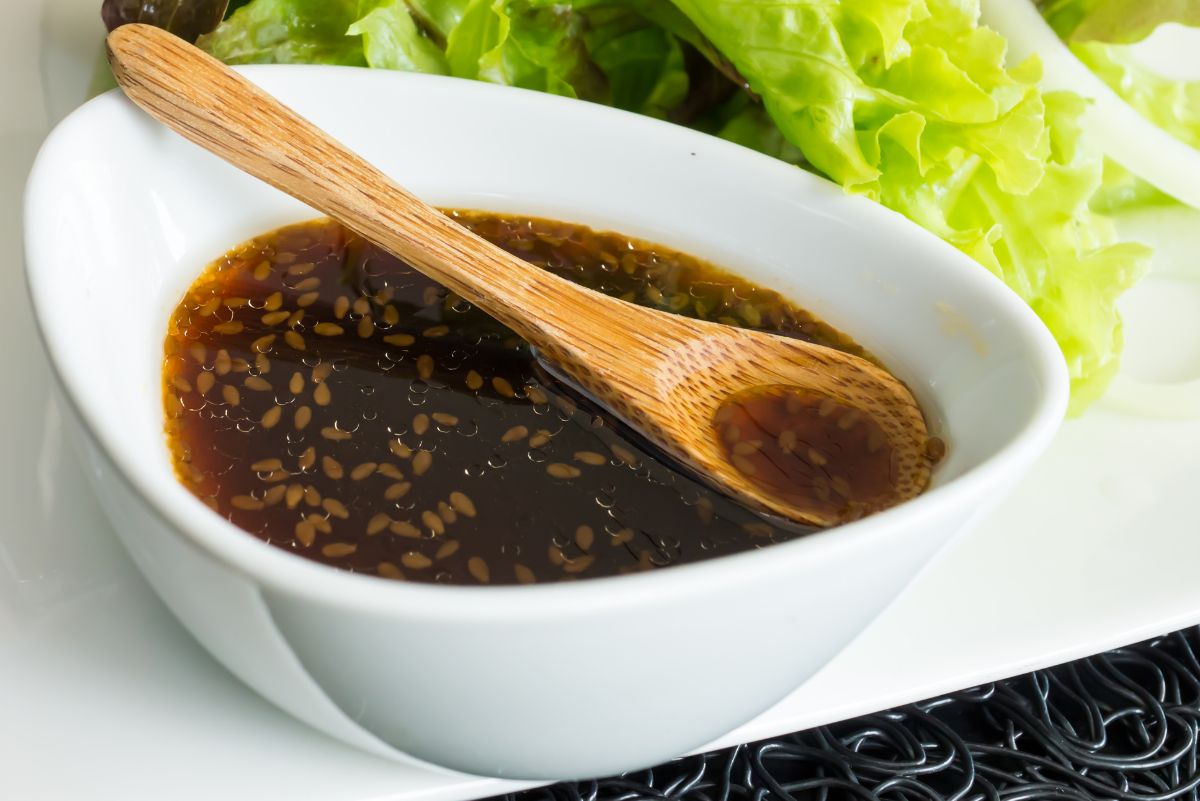 Japanese sesame soy salad dressing in a white bowl with sesame seeds, a wooden spoon, and fresh green lettuce on a white plate.