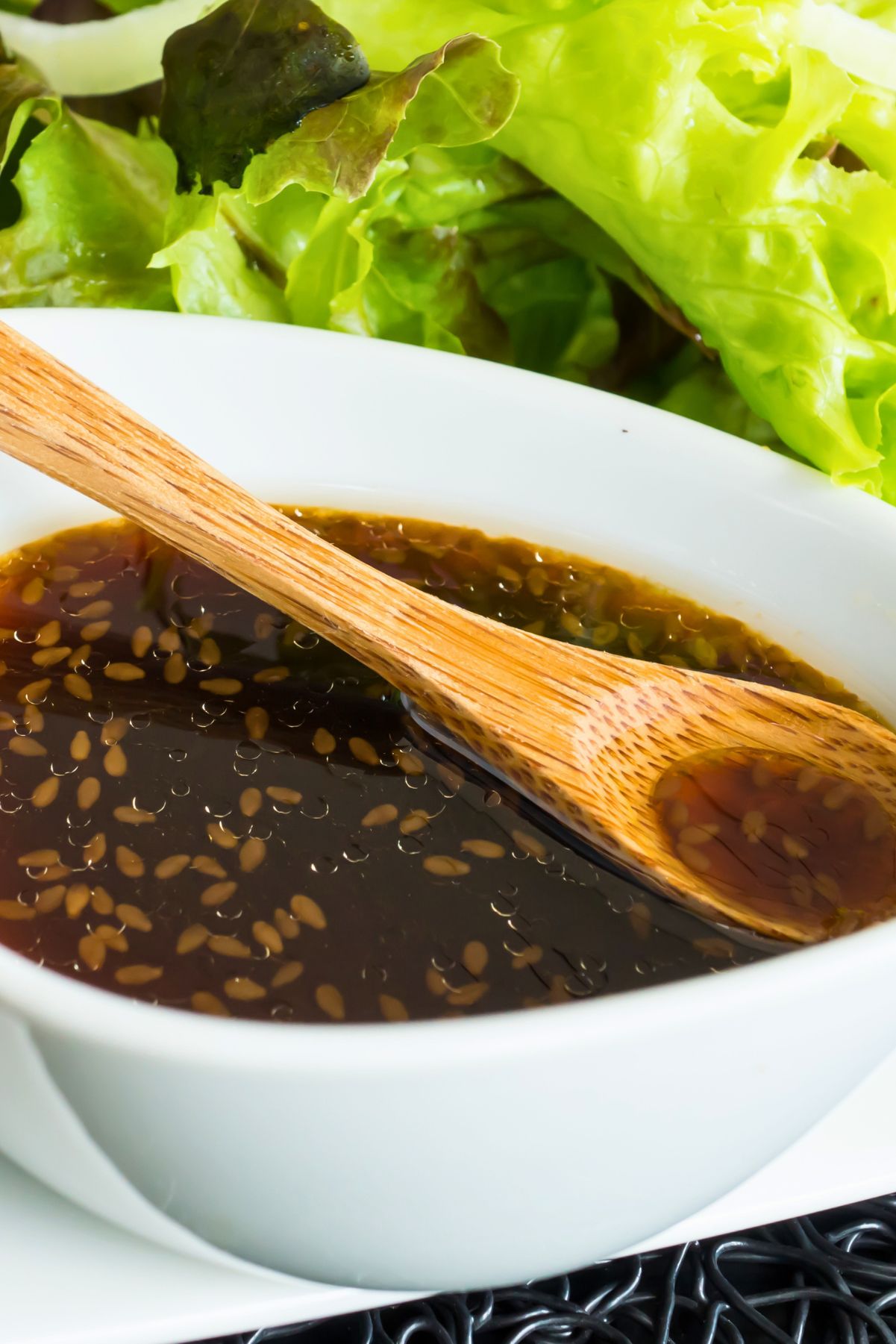 Japanese sesame soy salad dressing in a white bowl with sesame seeds, a wooden spoon, and fresh green lettuce in the background.