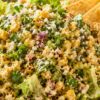 Mexican street corn chopped salad with romaine lettuce, roasted corn, red onion, cilantro, cotija cheese, chili seasoning, creamy dressing, and tortilla chips in a bowl.