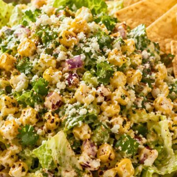 Mexican street corn chopped salad with romaine lettuce, roasted corn, red onion, cilantro, cotija cheese, chili seasoning, creamy dressing, and tortilla chips in a bowl.