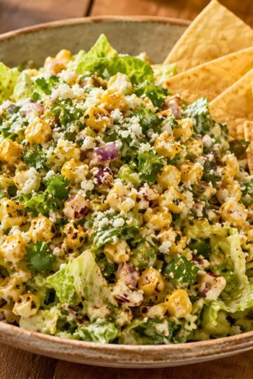 Mexican street corn chopped salad with romaine lettuce, roasted corn, red onion, cilantro, cotija cheese, chili seasoning, creamy dressing, and tortilla chips in a bowl.