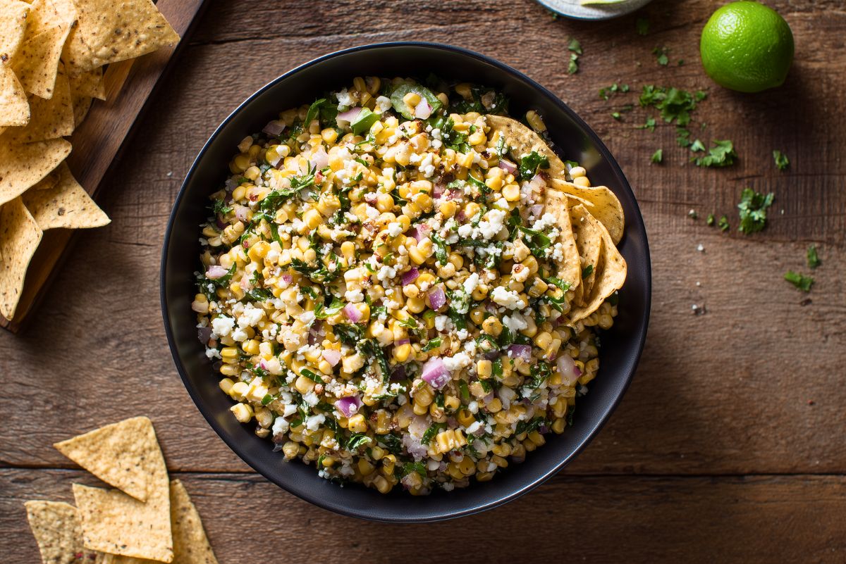Mexican street corn salad with cotija cheese, red onion, cilantro, and chopped greens, served with tortilla chips and lime.