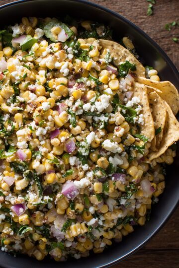 Mexican street corn chopped salad with corn, cotija cheese, red onion, cilantro, chopped greens, and tortilla chips in a black bowl.