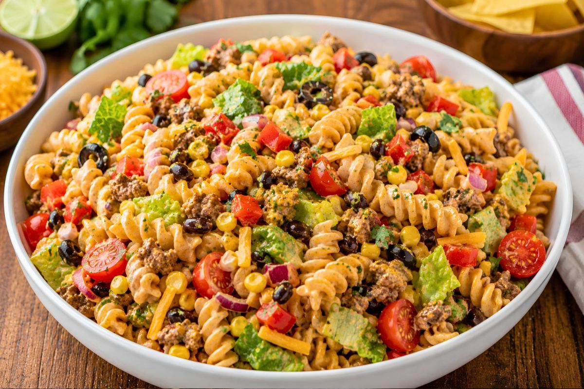 Southwest taco pasta salad in a white bowl with rotini, seasoned ground beef, corn, black beans, cherry tomatoes, romaine, red onion, cheddar cheese, cilantro, and creamy taco dressing on a wooden table.