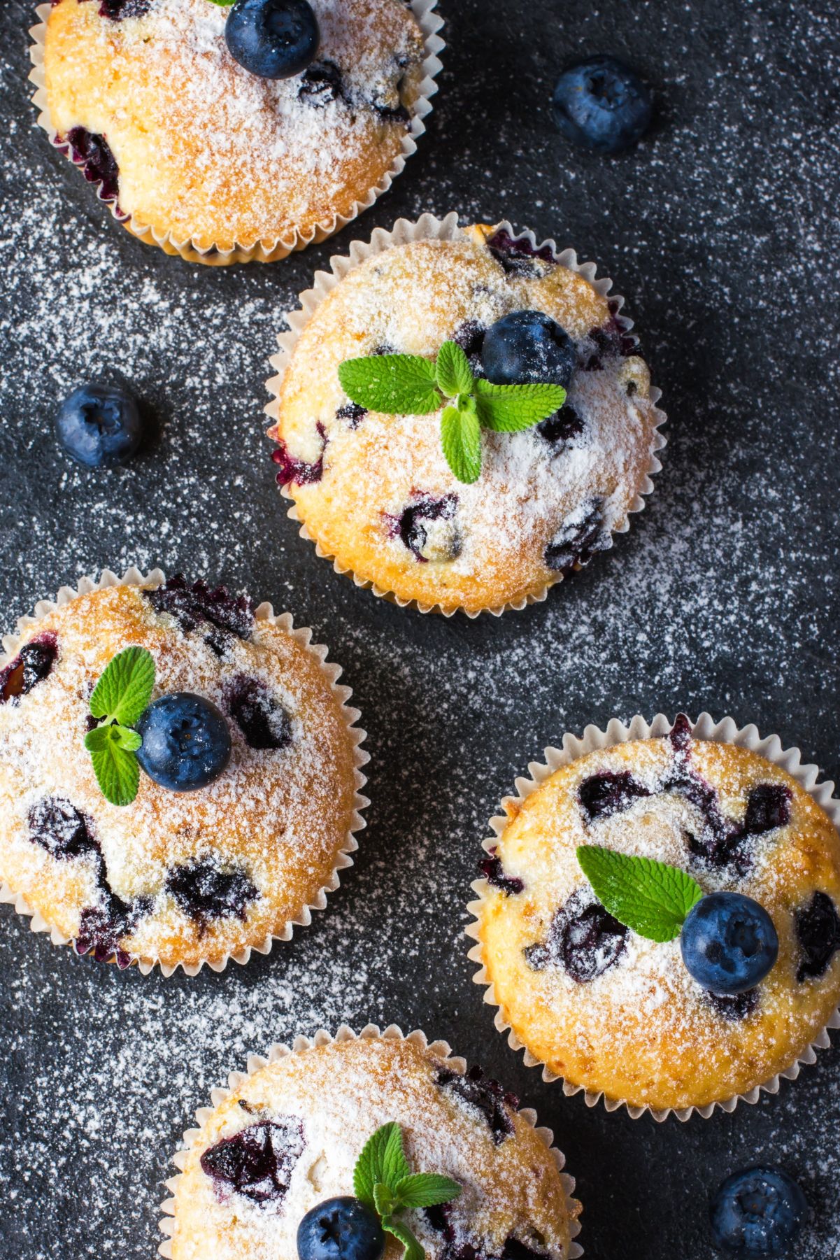 Air fryer blueberry muffins in paper liners, dusted with powdered sugar and topped with fresh blueberries and mint leaves on a dark surface with scattered blueberries.