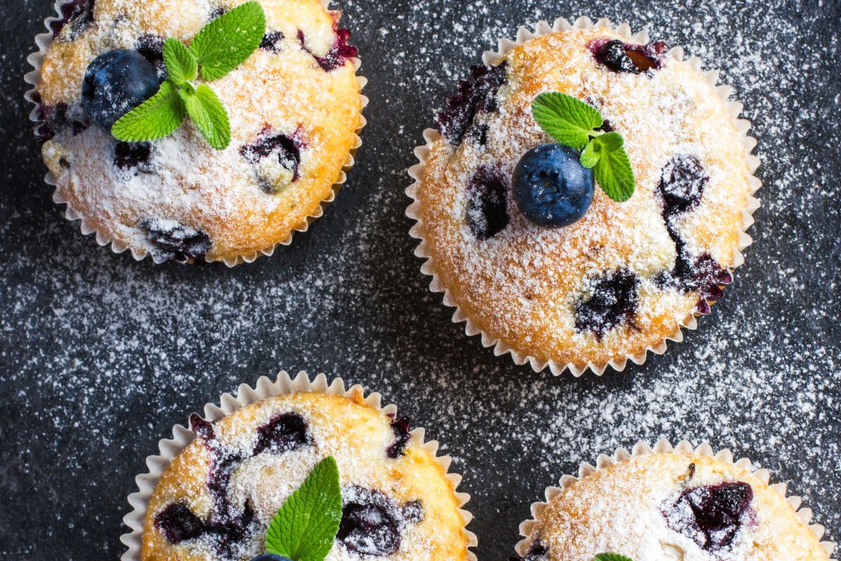 Air fryer blueberry muffins in paper liners, dusted with powdered sugar and topped with fresh blueberries and mint on a dark surface.