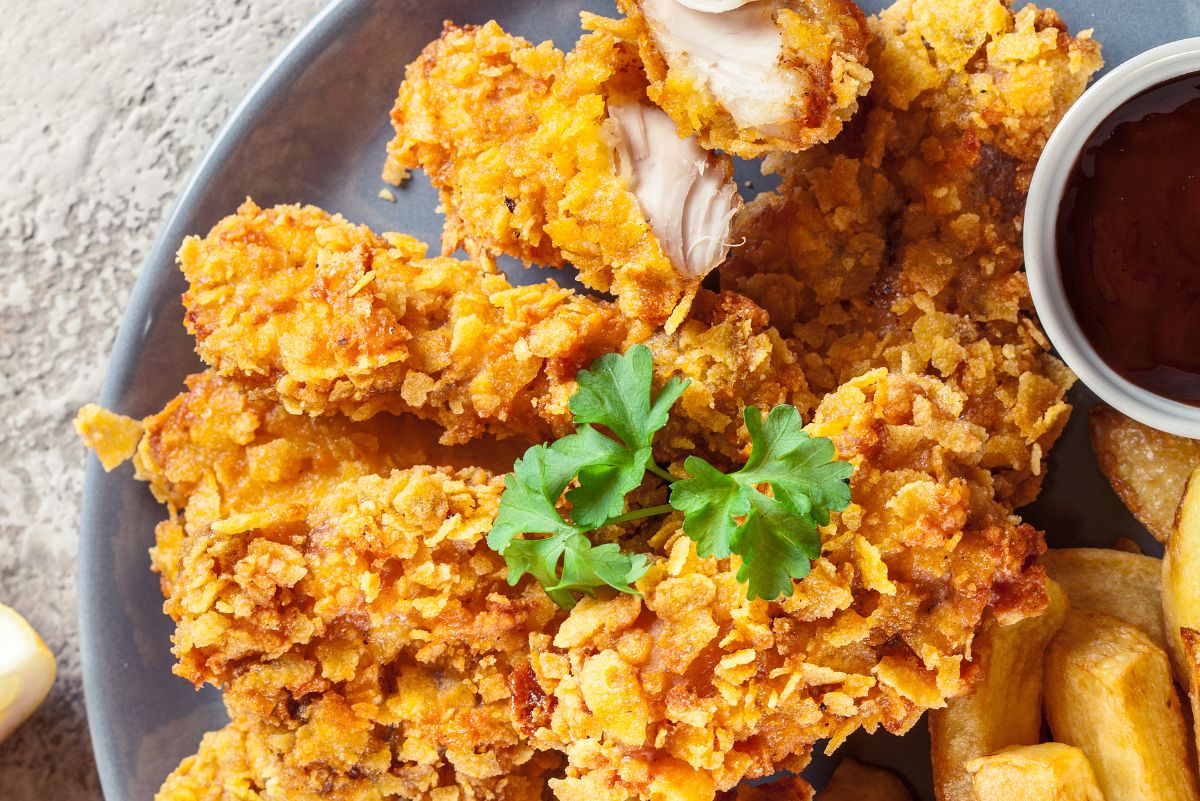 Southern oven-fried buttermilk chicken with a golden cornflake crust, served on a gray plate with thick-cut fries, parsley garnish, and a small bowl of dark dipping sauce.