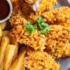 Southern oven-fried buttermilk chicken with a golden cornflake coating, served on a gray plate with thick-cut fries, parsley garnish, and a small bowl of dark dipping sauce.