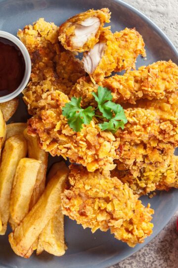 Southern oven-fried buttermilk chicken with a golden cornflake coating, served on a gray plate with thick-cut fries, parsley garnish, and a small bowl of dark dipping sauce.