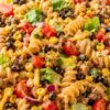 Southwest taco pasta salad with rotini, seasoned ground beef, corn, black beans, cherry tomatoes, romaine, red onion, cheddar, cilantro, and creamy taco dressing in a white bowl.