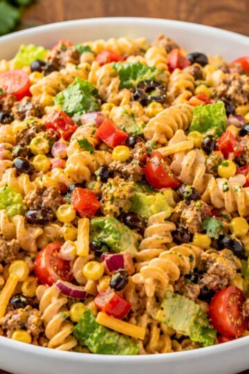 Southwest taco pasta salad with rotini, seasoned ground beef, corn, black beans, cherry tomatoes, romaine, red onion, cheddar, cilantro, and creamy taco dressing in a white bowl.