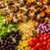 High protein taco chicken sweet potato bowl with grilled chicken, roasted sweet potatoes, romaine lettuce, black beans, corn, shredded cheese, red onion, cherry tomatoes, red bell pepper, cilantro, and creamy dressing.