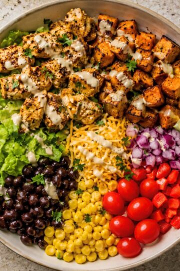 High protein taco chicken sweet potato bowl with grilled chicken, roasted sweet potatoes, romaine lettuce, black beans, corn, shredded cheese, red onion, cherry tomatoes, red bell pepper, cilantro, and creamy dressing.