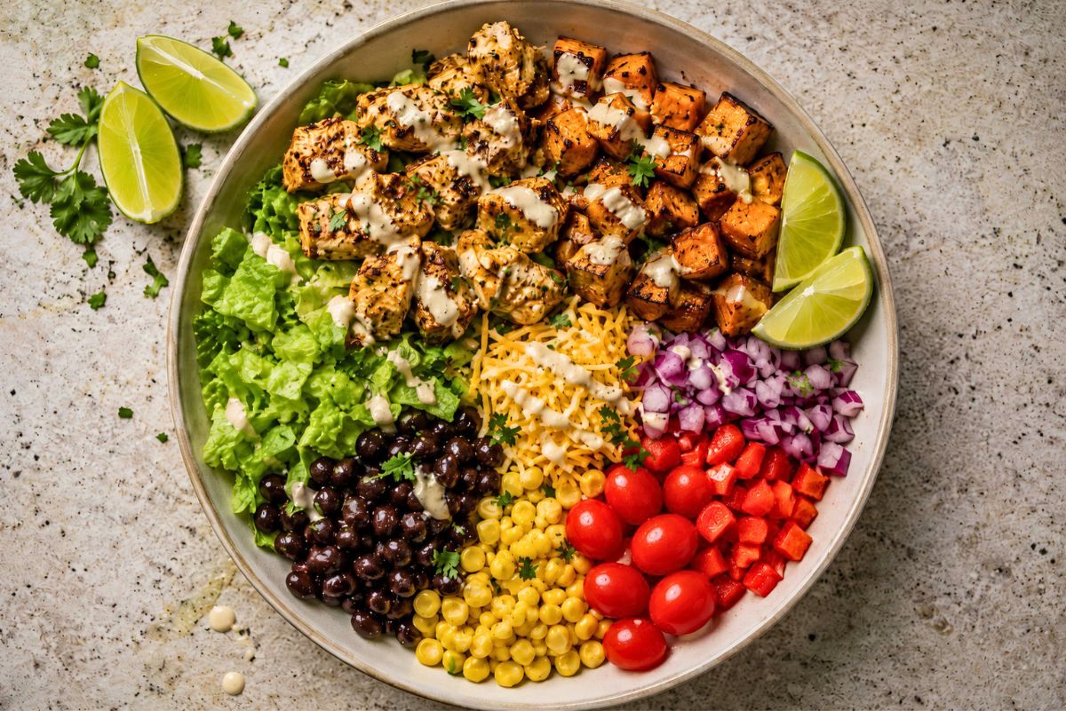 High protein taco chicken sweet potato bowl with grilled chicken, roasted sweet potatoes, romaine lettuce, black beans, corn, shredded cheese, red onion, cherry tomatoes, red bell pepper, lime wedges, cilantro, and creamy dressing.