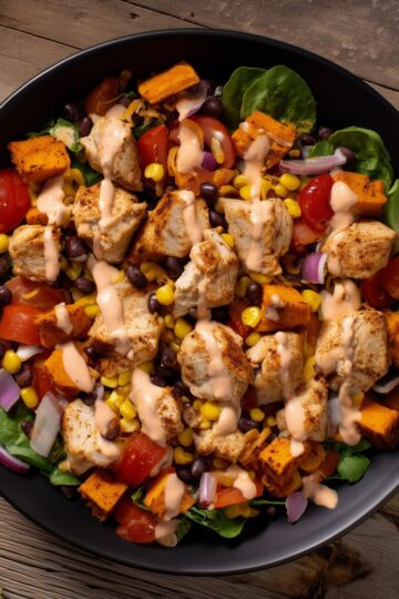 Close-up of a chicken taco sweet potato protein bowl with black beans, corn, tomatoes, red onion, roasted sweet potatoes, and creamy dressing.