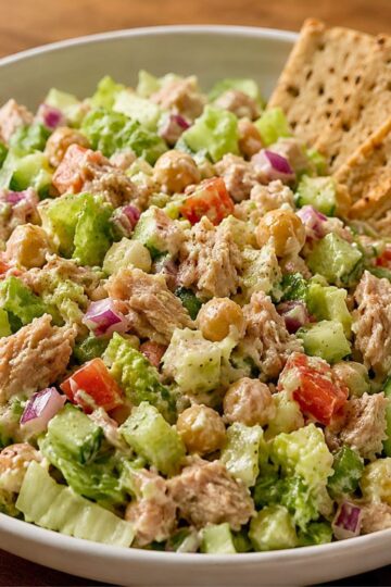 Tuna chopped salad with chickpeas, romaine lettuce, cucumber, red onion, tomato, and creamy dressing in a white bowl, served with crisp crackers on the side.