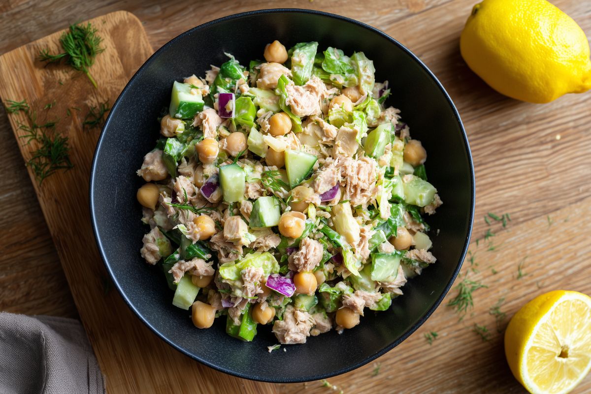 Tuna chickpea chopped salad with cucumbers, romaine, red onion, dill, and creamy dressing beside fresh lemons on a wooden table.