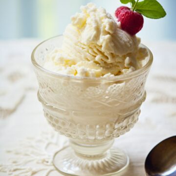 Vanilla frozen yogurt scooped into a vintage glass dessert cup, topped with a fresh raspberry, green leaves, and tiny white flowers. A silver spoon rests on a white lace tablecloth beside the creamy frozen dessert.