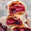 Three air fryer cherry pie dessert rolls stacked on a wooden board, showing crisp golden wrappers and glossy homemade cherry filling. Whole and halved cherries sit beside the rolls with a light dusting of powdered sugar.