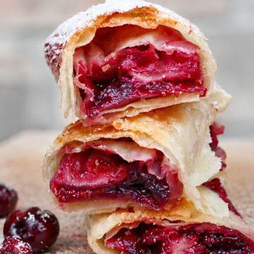 Three air fryer cherry pie dessert rolls stacked on a wooden board, showing crisp golden wrappers and glossy homemade cherry filling. Whole and halved cherries sit beside the rolls with a light dusting of powdered sugar.