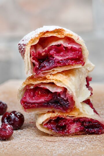 Three air fryer cherry pie dessert rolls stacked on a wooden board, showing crisp golden wrappers and glossy homemade cherry filling. Whole and halved cherries sit beside the rolls with a light dusting of powdered sugar.