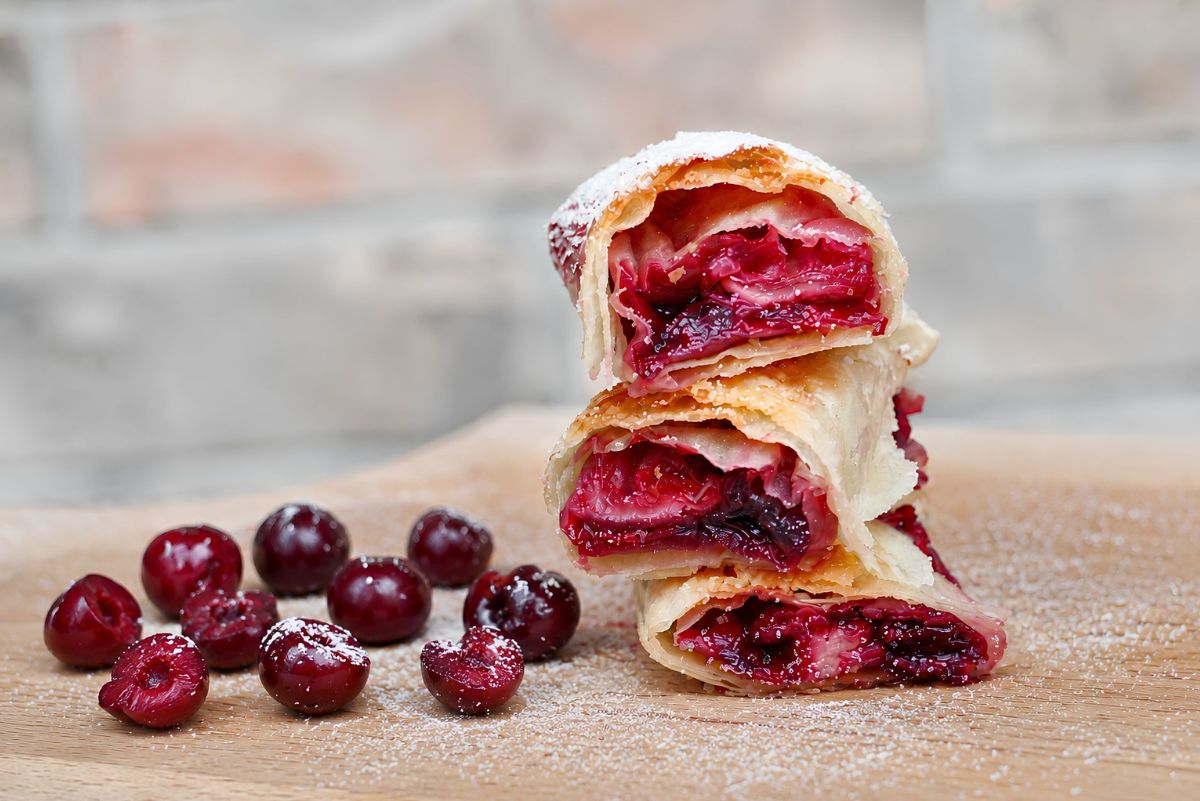 Three crispy air fryer cherry pie dessert rolls stacked on a wooden board, showing bright red cherry filling inside golden wrappers. Whole and halved cherries sit beside the rolls with a light dusting of powdered sugar.