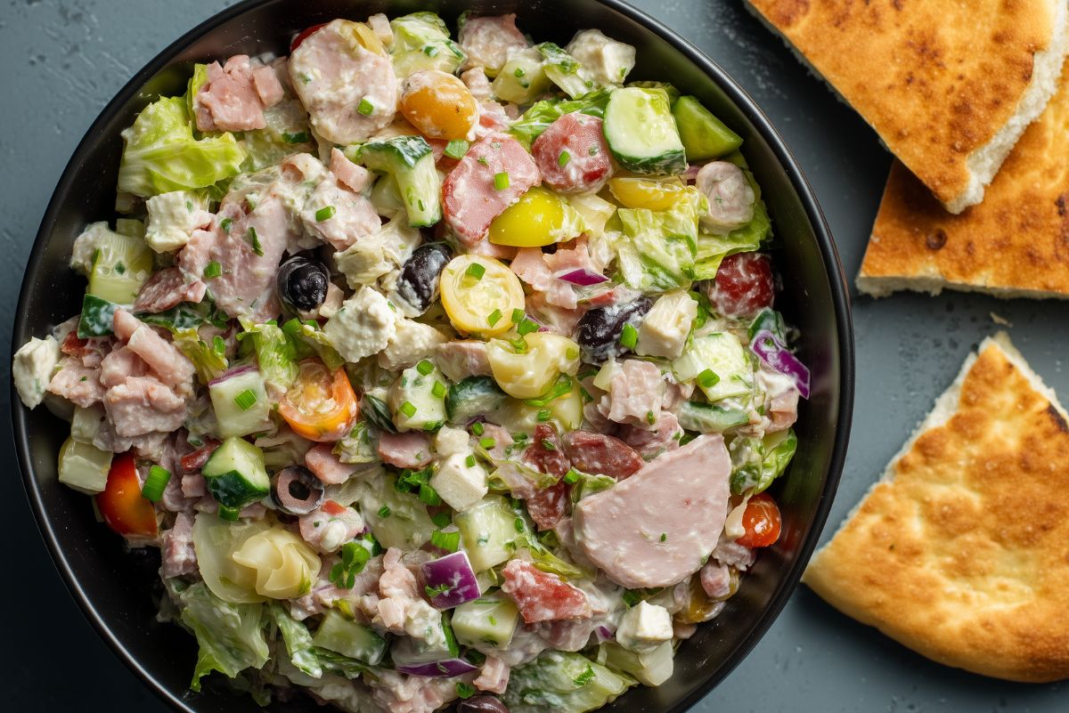 Overhead view of antipasto grinder chopped salad with deli meats, lettuce, cucumbers, tomatoes, olives, cheese, and creamy dressing, served with toasted bread.