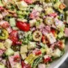 Antipasto grinder chopped salad with romaine lettuce, ham, salami, provolone, cherry tomatoes, cucumbers, artichoke hearts, roasted red peppers, banana peppers, black and green olives, and creamy dressing in a white bowl.